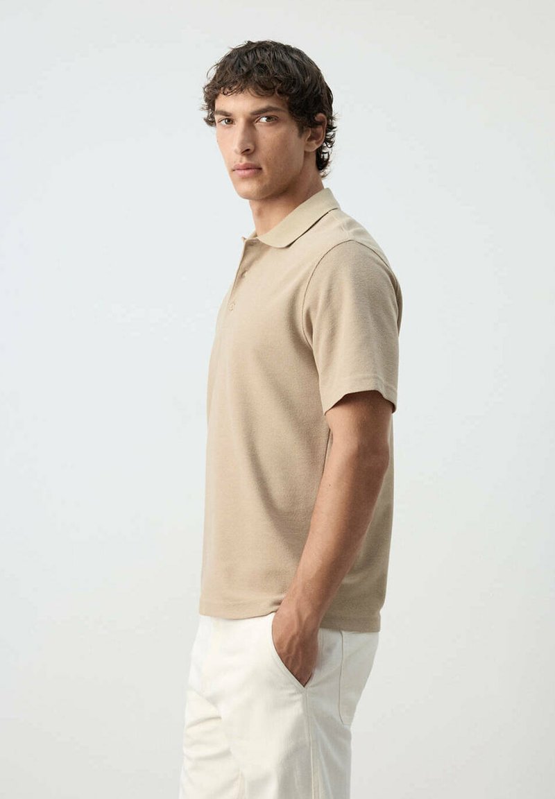 Hombre con polo beige de manga corta y pantalones blancos de pie de lado con una mano en el bolsillo contra un fondo liso y claro.