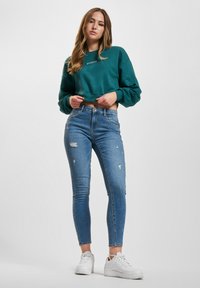 Grønn cropped sweatshirt med teksten "avenue", kombinert med slitte blå skinny jeans og hvite sneakers. Glatt tekstur, avslappet passform.