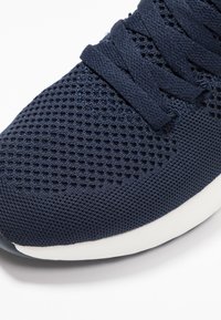 Granatowe siatkowe sneakersy z teksturowanym wierzchem, płaskimi sznurówkami i białą podeszwą. Posiadają perforowany wzór zapewniający wentylację i elastyczność.