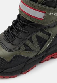 Zapatilla alta en negro y verde oliva con parte superior de cuero texturizado y malla, suela roja y una correa ajustable en gris y rojo.
