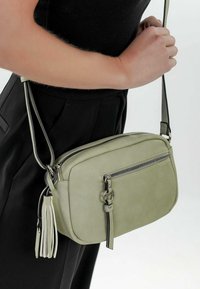 Borsa a tracolla verde in pelle con chiusura a zip, tracolla regolabile e dettaglio di frange decorative. Texture liscia e forma rotonda.