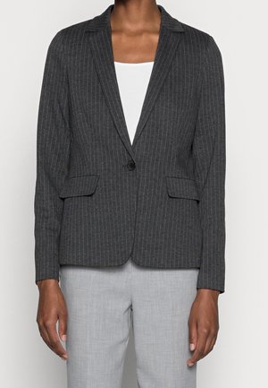Mørkegrå blazer med nålestriber og én knap, båret over en hvid top og lysegrå bukser, på en person, der står foran en ensfarvet baggrund.