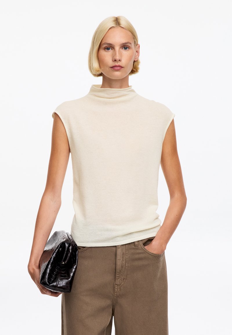 Femme aux cheveux blonds courts portant un haut crème sans manches et un pantalon marron, tenant une pochette noire texturée dans une main, l’autre main dans la poche.