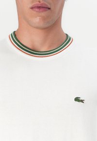 Hvit strikkegenser med ribbestrikket krage som har grønne, hvite og oransje striper; liten grønn krokodillelogo på venstre bryst.