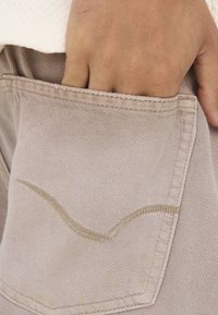 Pantalones de mezclilla beige claro con un diseño de bolsillo trasero cosido y una mano parcialmente metida en el bolsillo, mostrando detalles de textura y costuras.