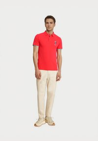 Rde poloko s ovratnikom, modrimi detajli na rokavih in majhno vezeno obliko. V kombinaciji s bež hlačami in svetlimi supergami.