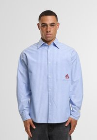 Light blue striped long-sleeve shirt made of cotton, featuring a chest pocket and red embroidered logo on the pocket. Plain collar, button-front design.