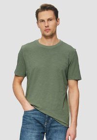 Oliwkowa koszulka z krótkim rękawem z miękkiego materiału z okrągłym dekoltem, zestawiona z niebieskimi dżinsami. Posiada luźny krój i prosty design.