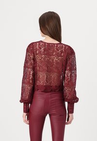 Burgundy lace top with long, puffed sleeves and floral patterns, featuring a sheer back and scalloped hem, paired with matching fitted pants.