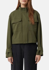 Frau in einer olivgrünen, taillierten Jacke mit vorderen Klappen-Taschen und schwarzen, weit geschnittenen Hosen, die vor einem schlichten Hintergrund steht.