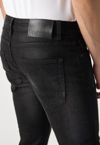 Gros plan sur une personne portant un jean noir ajusté avec une poche arrière et un patch en cuir noir marqué "INDICODE" à la ceinture.