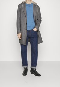Cappotto grigio di lana con cappuccio, camicia blu, jeans in denim scuro e stivali neri alla caviglia. Il cappotto ha bottoni e una vestibilità lunga, i jeans sono risvoltati alle caviglie.