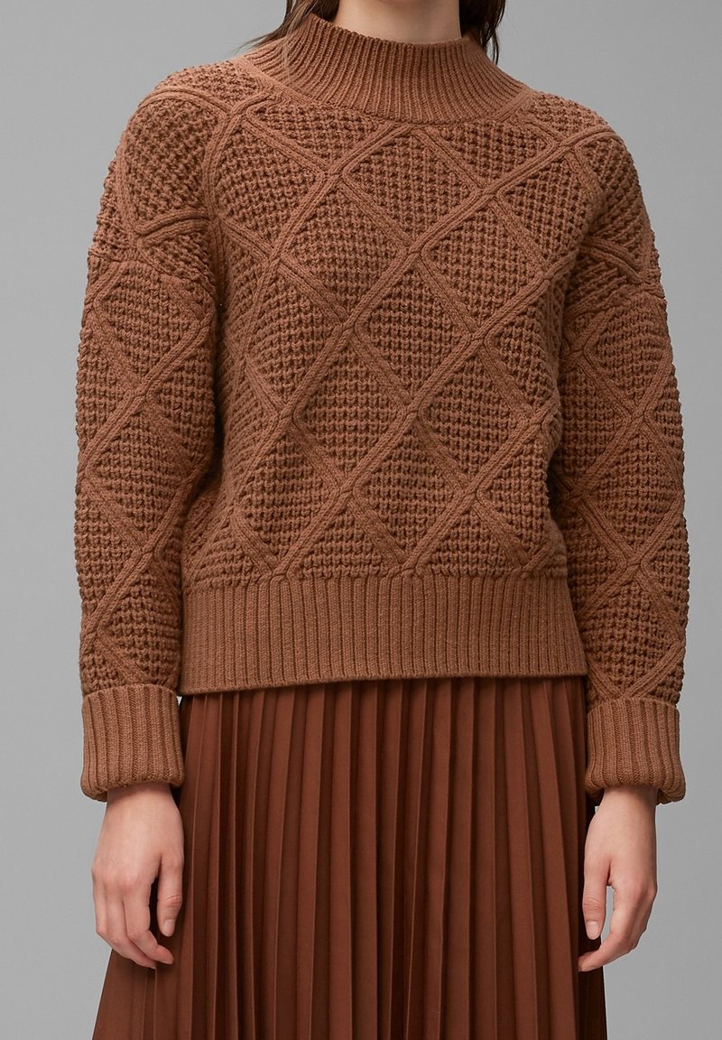 Pull en maille grosse torsade marron avec motif en losanges, poignets et ourlet côtelés, porté avec une jupe plissée marron assortie.