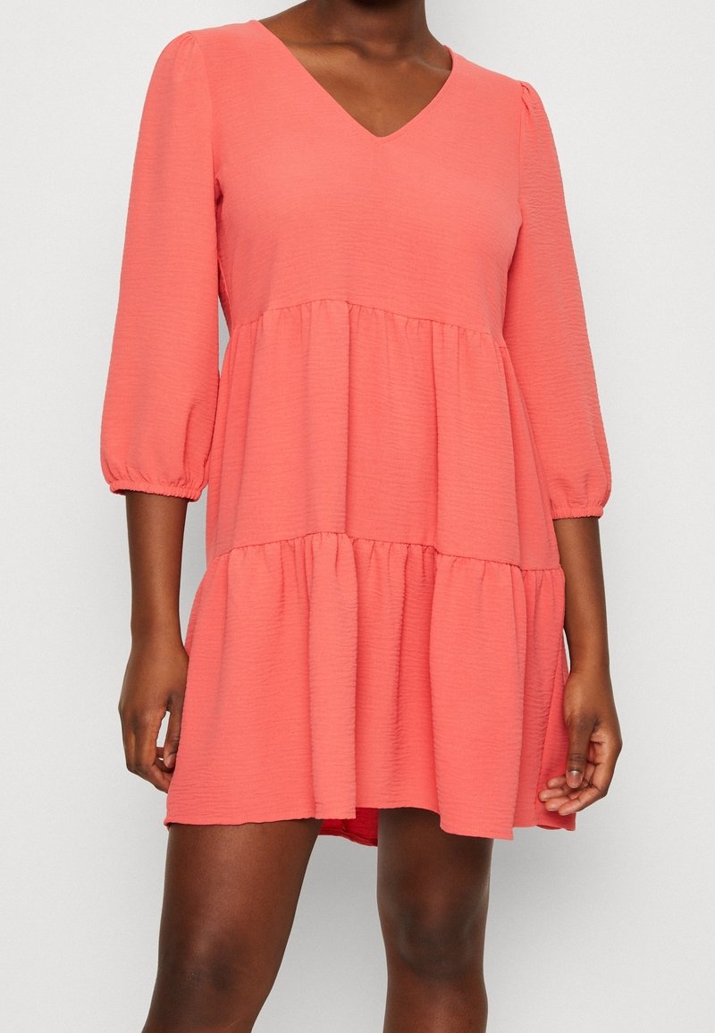 Femme portant une mini-robe corail rose à étages avec manches bouffantes trois-quarts et encolure en V sur un fond uni.