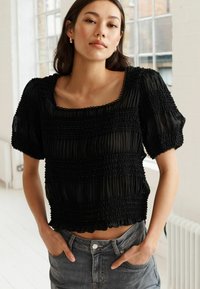 Černý zkrácený top s texturovaným smockingem, který má krátké nabírané rukávy a čtvercový výstřih. Kombinován s džínovými šortkami na jasném pozadí.