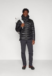 Giacca a vento nera con colletto alto, design elegante e texture lucida. Abbinata a pantaloni scuri e scarponi da trekking robusti. Zaino bianco.