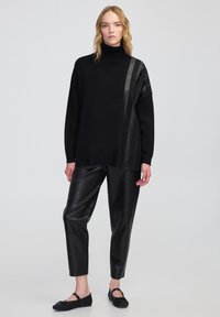 Kobieta w czarnej golfie z skórzanymi paskami, czarnych skórzanych spodniach i czarnych balerinach Mary Jane, stojąca na jasnym tle.