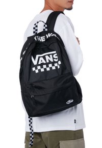 Zaino in tessuto nero con logo bianco "VANS" e spallacci a scacchi, dotato di una tasca frontale e spallacci imbottiti regolabili.