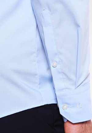 Poignet de chemise bleu clair à manches longues avec des boutons blancs et des boutonnières cousues, porté avec un pantalon foncé.