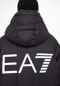 Melnā pūkainā jaka ar lielu balto "EA7" logotipu un svītrainām akcentiem aizmugurē, ar kapuci un quilted tekstūru.