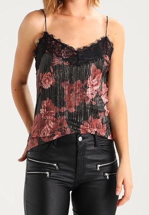 Femme portant un caraco noir à fleurs rouges avec des bordures en dentelle et des bretelles fines, ainsi qu'un pantalon en cuir noir avec plusieurs fermetures éclair à l'avant.