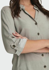 Camisa de botones de color gris claro con mangas enrolladas, que presenta un tejido texturizado y botones oscuros. El cuello está abierto, revelando un diseño casual.