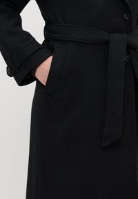 Melna vilnas mēteli ar jostu, kuram ir pogājamas piedurknes un sānu kabatas. Audums izskatās mīksts un teksturēts.