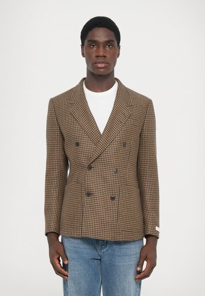 Brun houndstooth dobbeltknap blazer med sorte knapper, tekstureret stof, slag med notch og en tætsiddende pasform. Båret over en hvid skjorte.