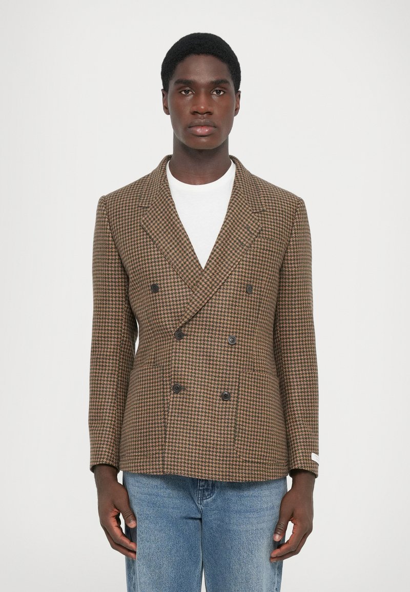 Brun houndstooth dubbelknäppt blazer med svarta knappar, texturerat tyg, slagkläppen och en figursydd design. Bärs över en vit skjorta.