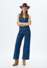 Outfit in denim composto da un top senza maniche con bottoni, jeans a gamba larga e stivali alla caviglia. Accentuato con un cappello marrone e collane a strati.