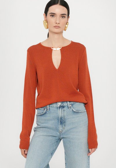 Rust-colored knit sweater with a keyhole neckline, long sleeves, and soft texture, paired with high-waisted light blue denim jeans.