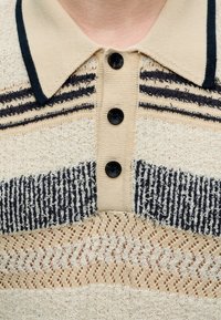 Polo en maille beige avec des rayures noires, des boutons noirs et un motif texturé sur la poitrine et le col.