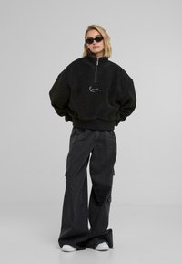 Pullover in pile nero con zip e colletto alto, caratterizzato da un logo bianco. Abbinato a pantaloni cargo in denim scuro e dalla vestibilità ampia e sneakers bianche.