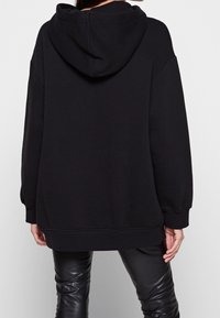 Persona che indossa una felpa nera oversize e pantaloni in pelle nera, vista di spalle su uno sfondo chiaro e semplice.