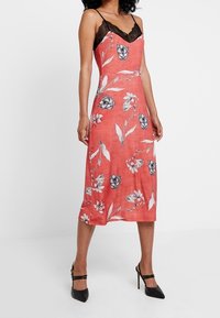 Robe florale rouge avec des accents en dentelle noire, bretelles spaghetti et une longueur mi-mollet. Présente une coupe ample et un motif floral vibrant sur le tissu.