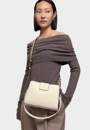 Donna che indossa un maglione taupe con spalle scoperte e pantaloni beige, porta una borsa a tracolla color crema con "TOUS" impresso sulla patta.