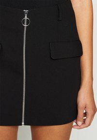 Gonna nera con zip frontale e tirante circolare, due tasche a patta e una texture di tessuto liscia. Progettata per una vestibilità sartoriale.