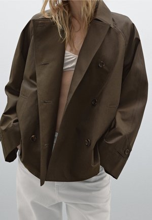 Personne portant une veste oversize double boutonnage marron foncé sur un crop top blanc et un pantalon blanc ample, mains dans les poches de la veste.