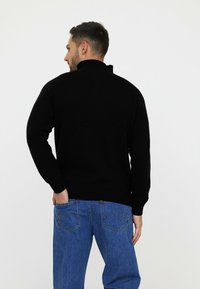 Pull noir en tricot avec un col montant, des poignets et un ourlet côtelés, porté avec un jean bleu montrant les détails de la poche arrière.