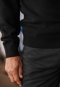 Juodas mezginys su raukinėmis rankovėmis, derinamas su tamsiomis tekstūruotomis kelnėmis. Dėmesys skiriamas rankai, ramiai besiilsinčiai ant liemens, kurią puošia žiedas.