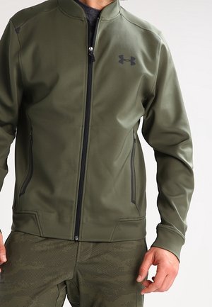Mann trägt eine olivgrüne Zip-up-Jacke mit schwarzem Reißverschluss und passende Hose im Tarnmuster, steht vor einem schlichten Hintergrund.