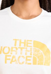 Camiseta de algodón blanca con un gran logo texturizado amarillo que incluye las palabras "THE NORTH FACE" y un diseño de medio círculo.