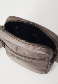 Bolso cruzado de tela marrón con un cierre superior con cremallera, superficie texturizada y un bolsillo frontal que muestra el logotipo "BOSS". El interior es negro.