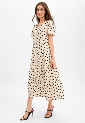 Donna che indossa un vestito midi beige con pois neri, bottoni frontali, maniche corte e sandali neri con tacco alto, in piedi su sfondo bianco.