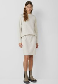 Roomkleurige gebreide trui en bijpassende rok, voorzien van een losse pasvorm en textuur. Gecombineerd met grijze enkelhoge laarzen, geplaatst tegen een neutrale achtergrond.