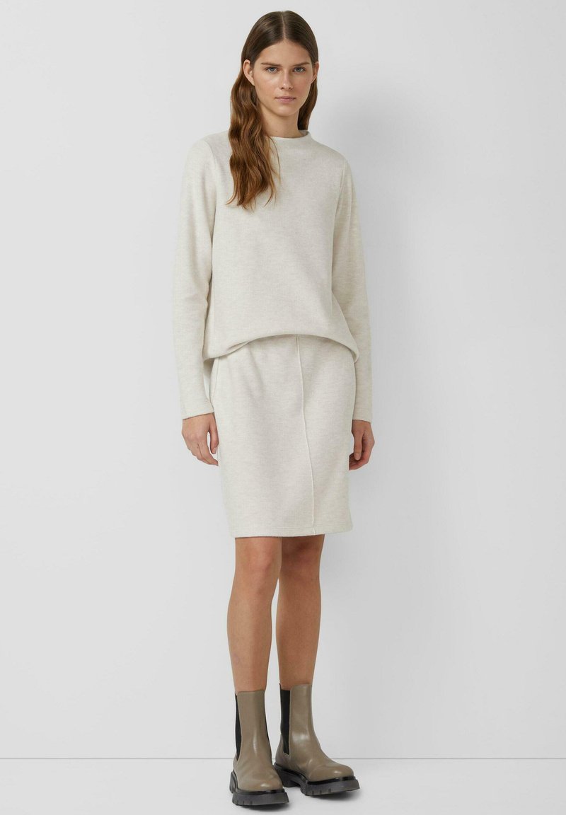 Roomkleurige gebreide trui en bijpassende rok, voorzien van een losse pasvorm en textuur. Gecombineerd met grijze enkelhoge laarzen, geplaatst tegen een neutrale achtergrond.