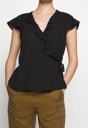 Femme portant un chemisier cache-cœur noir à manches courtes avec des volants et un pantalon marron moutarde, les mains dans les poches, debout devant un fond blanc.