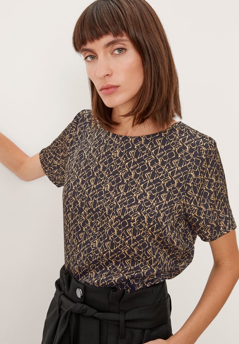 T-shirt nera a maniche corte con motivo astratto dorato, scollatura rotonda e vestibilità comoda. Abbinata a pantaloni neri a vita alta.