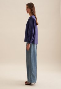 Maglietta viola a maniche lunghe con maniche strutturate, abbinata a jeans a gamba larga blu chiaro e sandali neri con tacco, mostrata di lato.