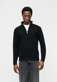 Νέος άνδρας που φοράει μαύρο πουλόβερ με φερμουάρ στο λαιμό πάνω από λευκό πουκάμισο και ξεθωριασμένο γκρι τζιν, στέκεται μπροστά σε απλό λευκό φόντο.